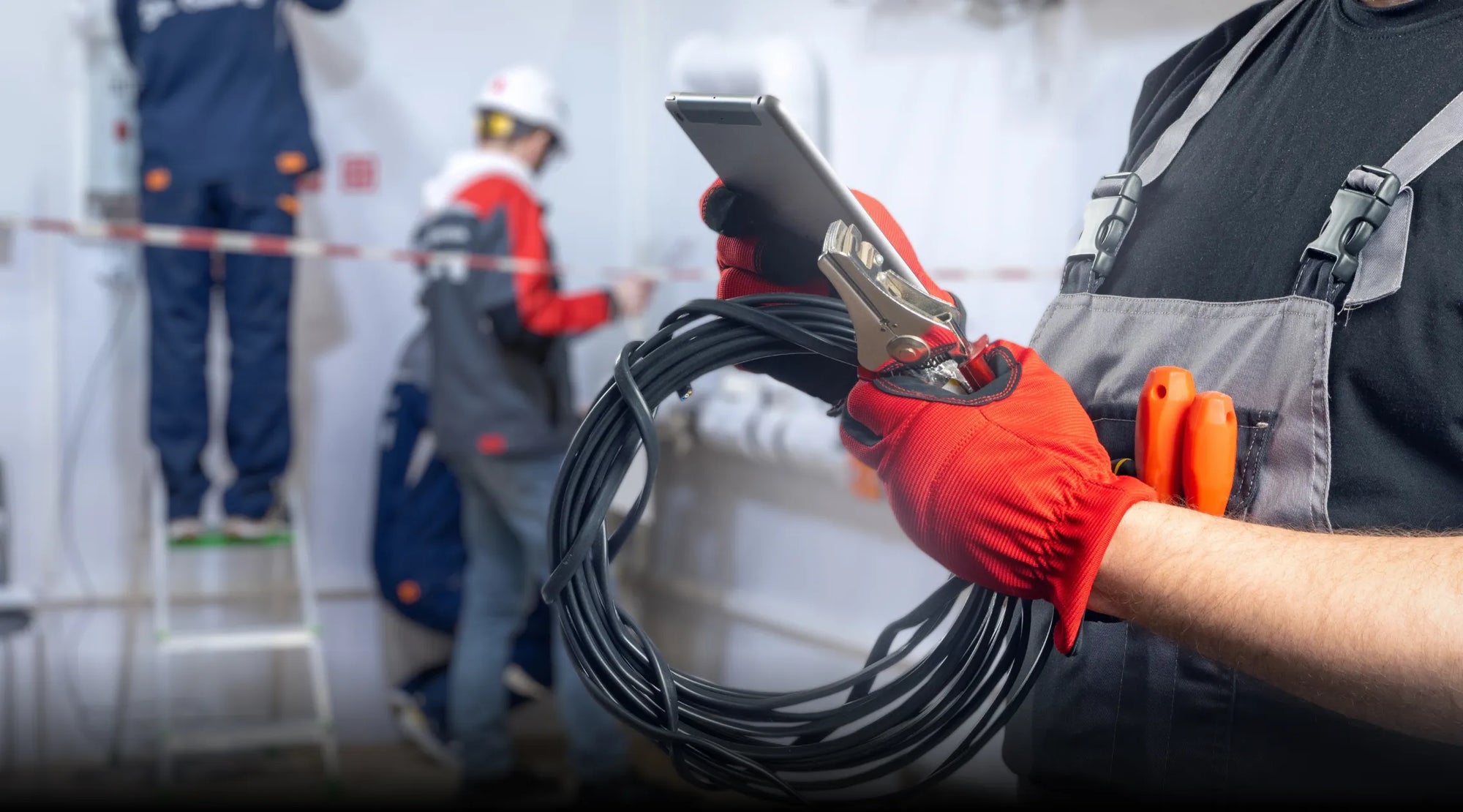 An installer in overalls and red gloves holds a coil of black cable and a tablet, with another worker on a ladder in the background, representing a professional installation.