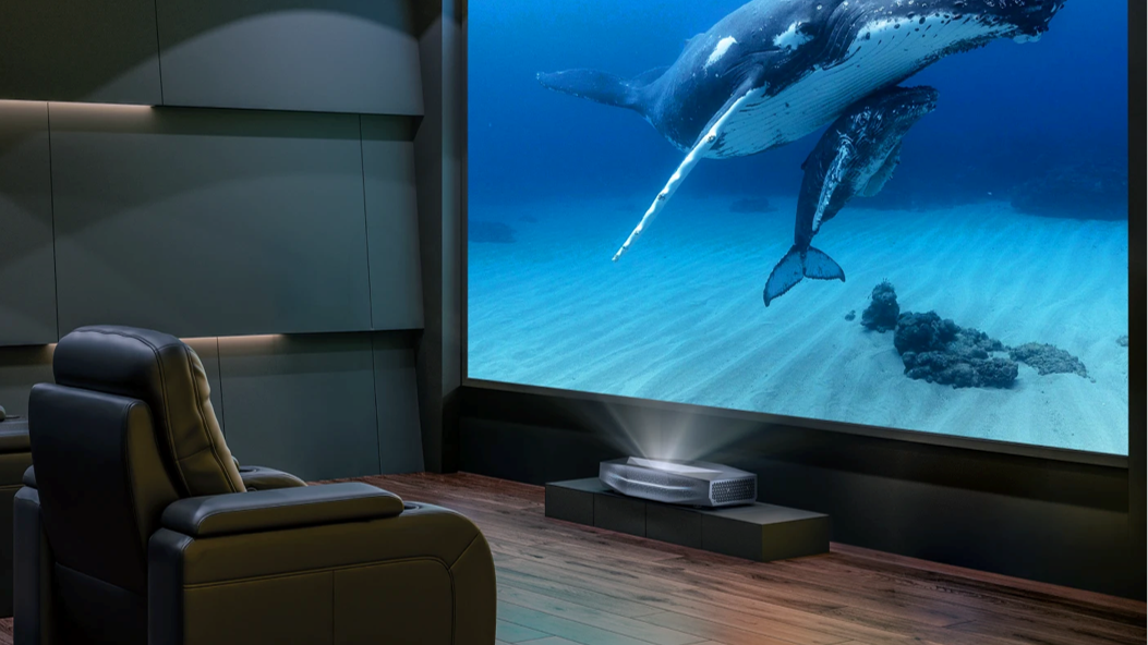 A sleek ultra short throw projector casts a vibrant, massive 4K image of humpback whales onto a screen in a dedicated, dark home theater with a leather recliner.
