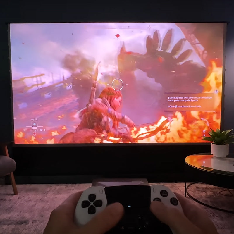 Cinematic+ ALR motorized floor rising projector screen used for gaming with a player holding a controller and playing an action game.