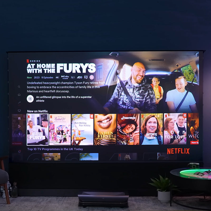 The Cinematic+ ALR motorized floor rising projector screen displays the Netflix interface and various program thumbnails.