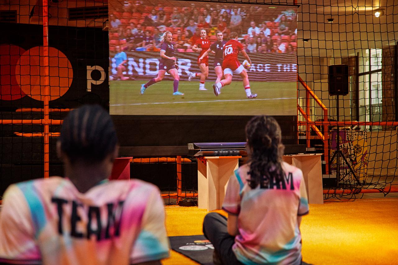 Two spectators, seen from behind, sit on mats watching a women's rugby match projected onto a large screen at the SOOO London event.
