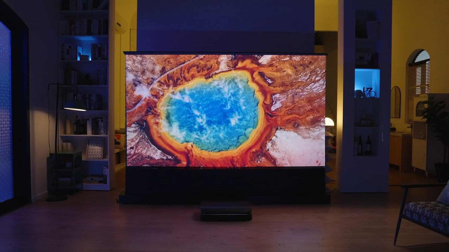 A stylish, dimly lit living room with a home cinema setup. An ultra-short-throw (UST) laser projector sits on the floor, projecting a massive, vivid 4K image of Yellowstone's Grand Prismatic Spring onto an ambient light rejecting (ALR) screen.