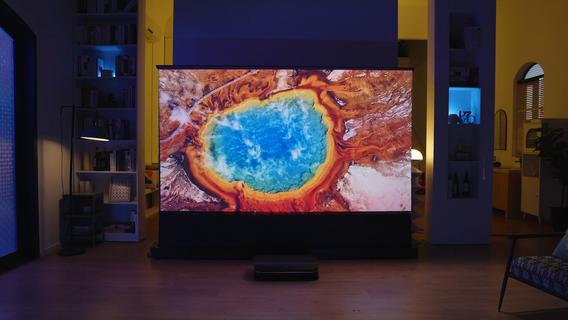 A stylish, dimly lit living room with a home cinema setup. An ultra-short-throw (UST) laser projector sits on the floor, projecting a massive, vivid 4K image of Yellowstone's Grand Prismatic Spring onto an ambient light rejecting (ALR) screen.