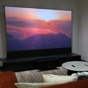AWOL Vision Laser TV in a home setting projects a sunrise over mountains on a large motorized screen, enhancing the ambient charm of a modern living room.